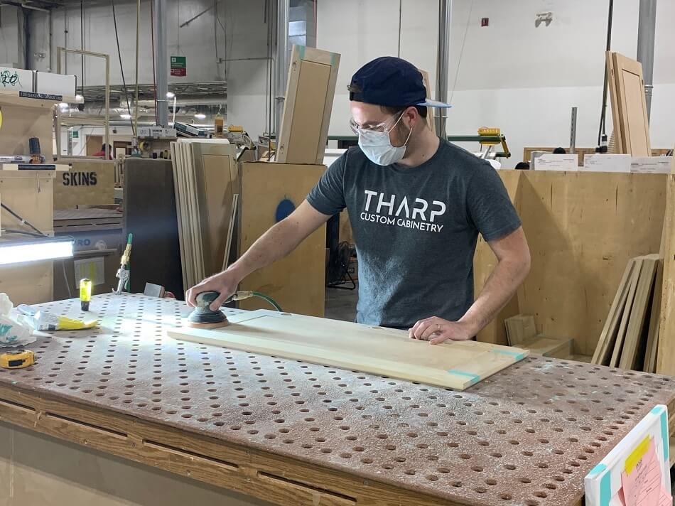 Man wearing a gray shirt that says Tharp Custom Cabinetry, a face mask and safety glasses while sanding a cabinet door in a wood workshop.