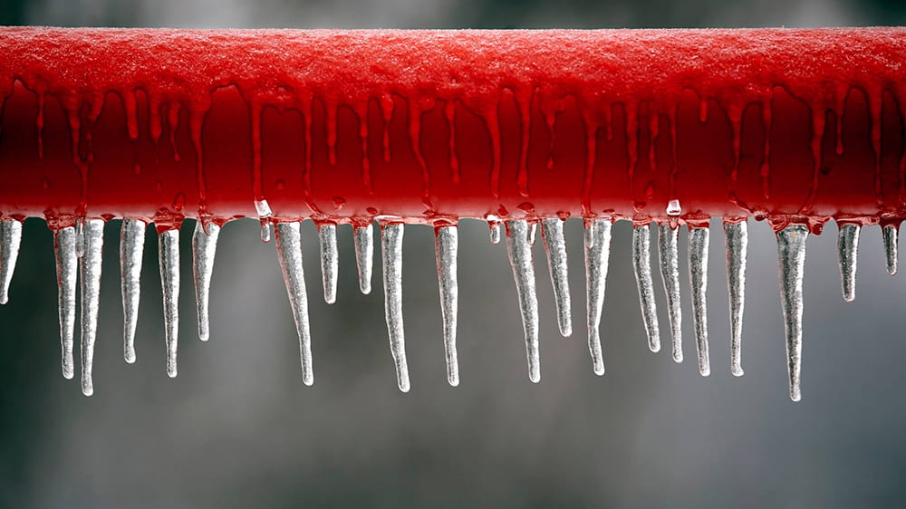 Red pipe frozen with a layer of ice and icicles frozen along the bottom edge after a winter storm in Texas.