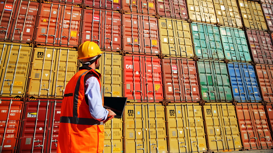 Dock worker reviewing stacks of large crates on a freight boat poses questions about current recession status.